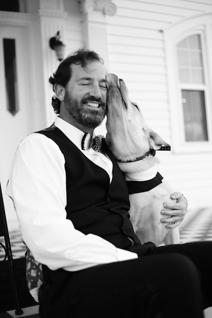 black and white photograph of groom sitting outside on the steps while his yellow lab dog licks his face -bisoux de lune meagan reily