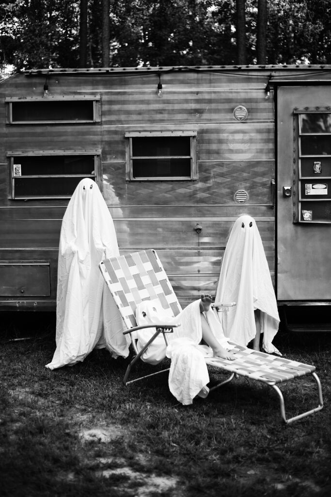 black and white photograph of a vintage airstream silver camper with children dressed in ghost costumes in a lawn chair