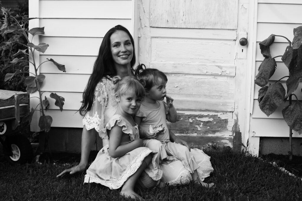 portrait of a stunning brunette model of a mother sitting in front of a worn down shed door surrounded by sun flowers while her two little girls in dresses sit in her lap smiling -bisoux de lune meagan reily