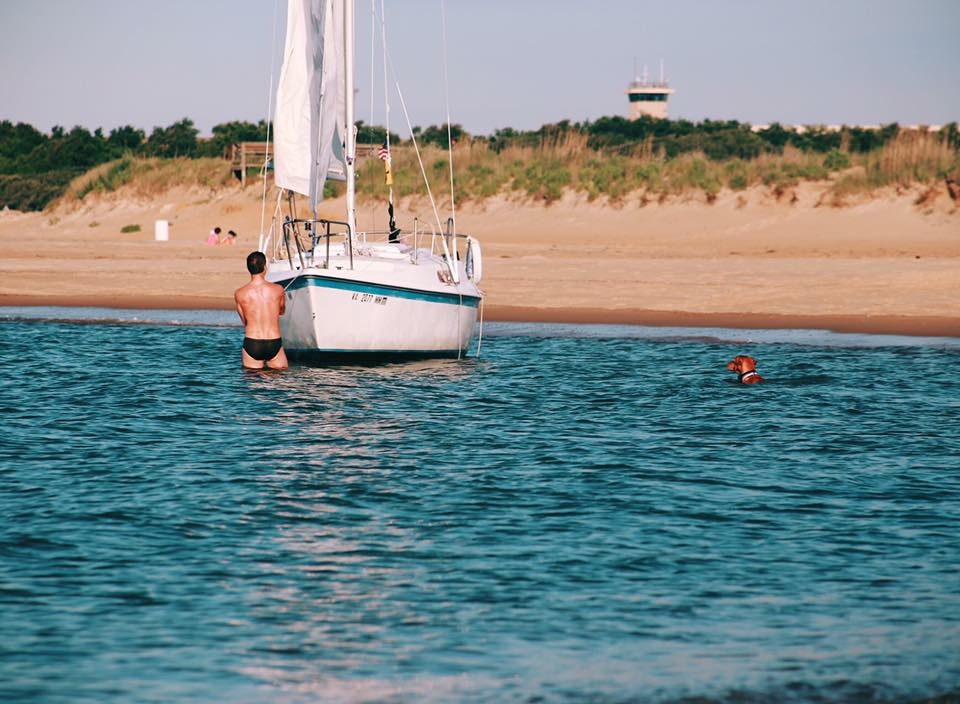 fine art photograph of a man in a black speedo with his golden retreiver in the water with him as he stands beside his sailboat and stares at the beach -bisoux de lune meagan reily