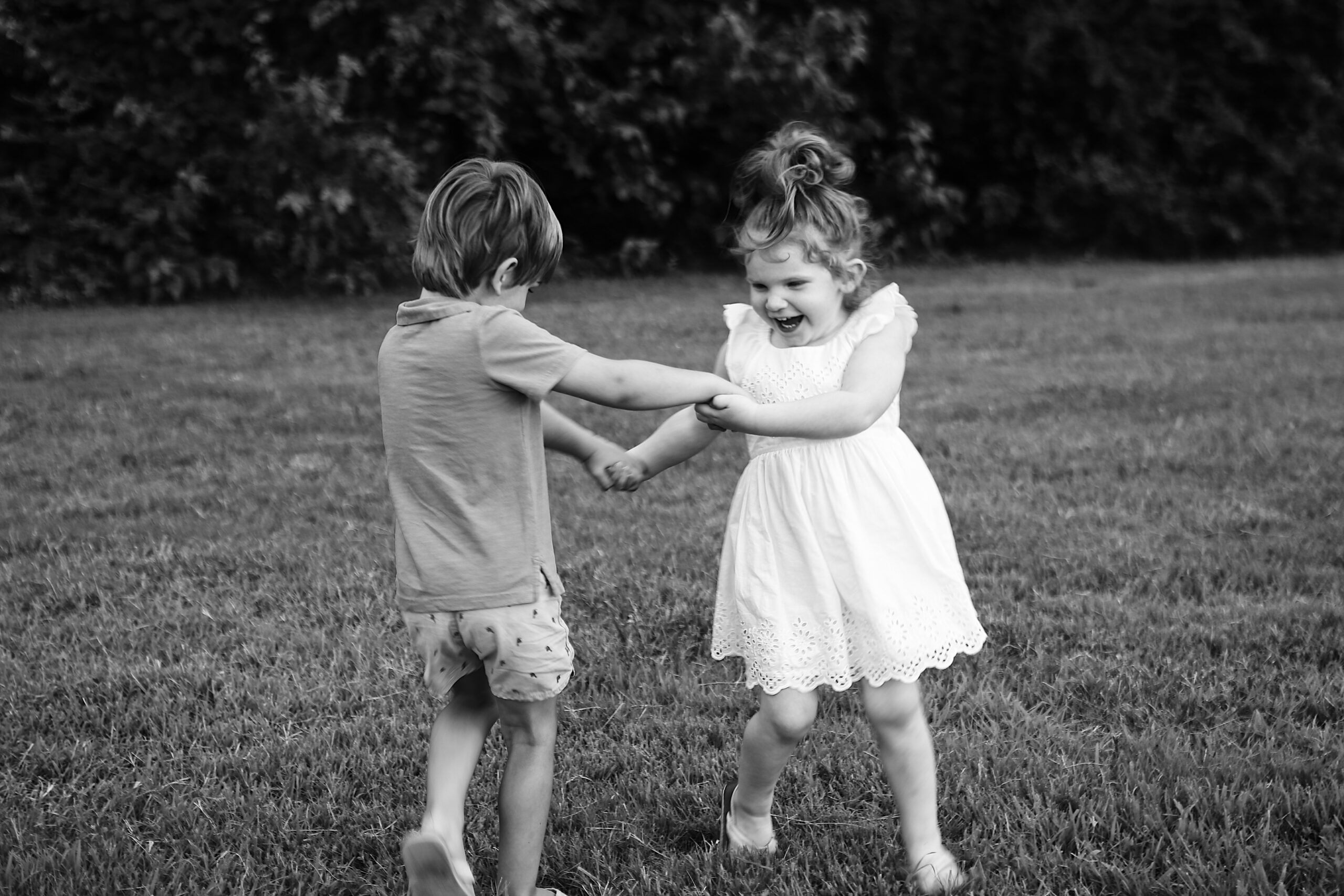 black and white portrait in a field of two young children no more than 5 years old holding hands and playing in a field while they laugh -bisoux de lune meagan reily