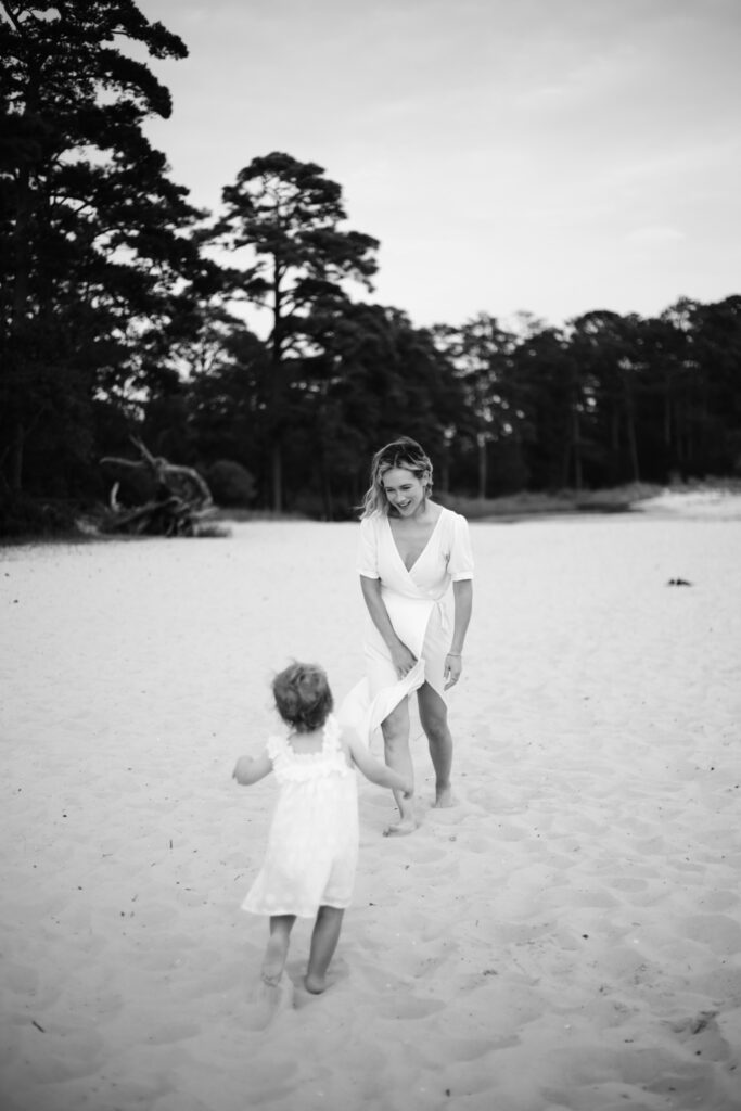 scenic beach shot of young beautiful mother and small baby running on the beach in virginia beach black and white photo with dark trees in the background the image is very whimsical and fun and sentimental there is water in the backgound -bisoux de lune meagan reily