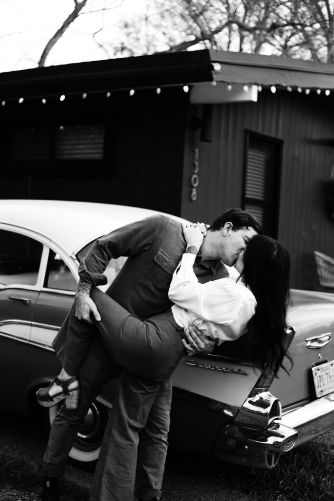 black and white holiday photograph in front of a black mid century home and a vintage antique chrysler or bel air of a couple dipping and kissing in vintage indie style clothing -bisoux de lune meagan reily