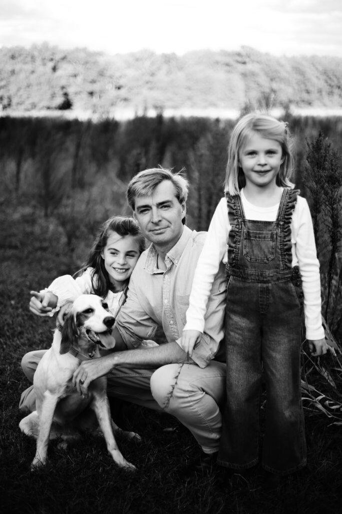 black and white photograph in the countryside of a grey haired father in a nice button down sitting in a field with his two young blonde daughters and their dog -bisoux de lune meagan reily