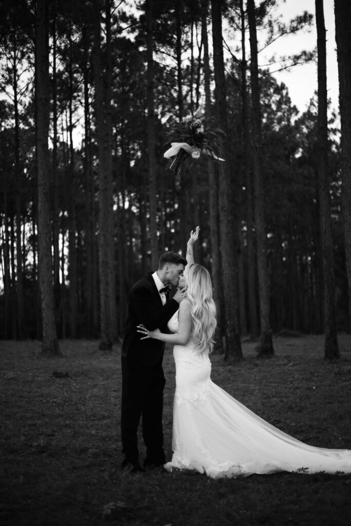 black and white portrait of a bride and groom throwing their wedding flowers in the woods while they kiss -bisoux de lune meagan reily