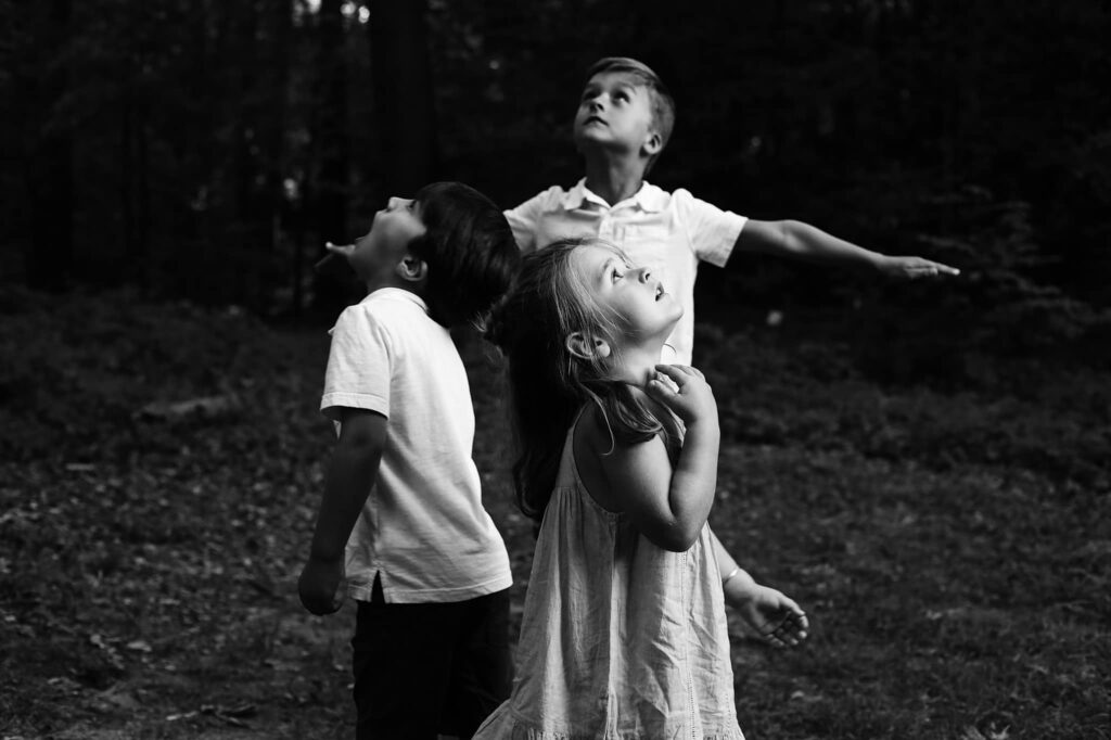 high contrast black and white photo of three children standing in a circle looking up at an airplane wihle one young boy holds his hands out like a plane -bisoux de lune meagan reily