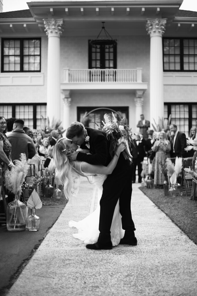 black and white photograph of a bride and groom dipping at the end of their aisle and kissing while onlookers clap and take photos in front of a huge southern mansion -bisoux de lune meagan reily