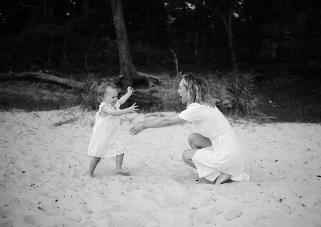 beach portrait of a young beautiful blonde mother in a white dress with her arms outstretched and her young two year old child in a white dress running into her arms with a smiles on their faces -bisoux de lune meagan reily