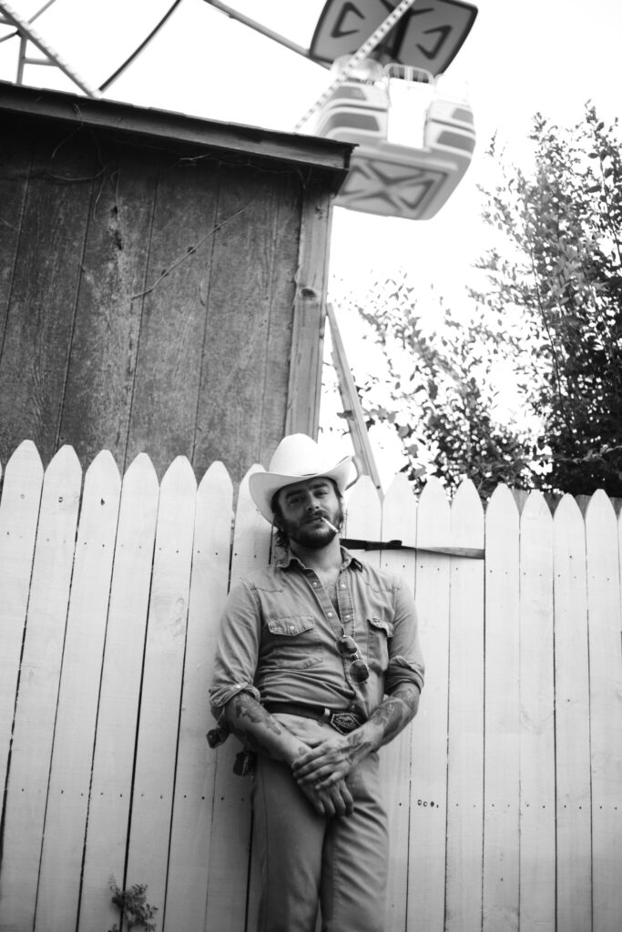 black and white photograph of a western style cowboy in a button down denim shirt and tweed pants standing in front of a fence with a cigarette in his mouth outside of a county fair with the ferris wheel in the background