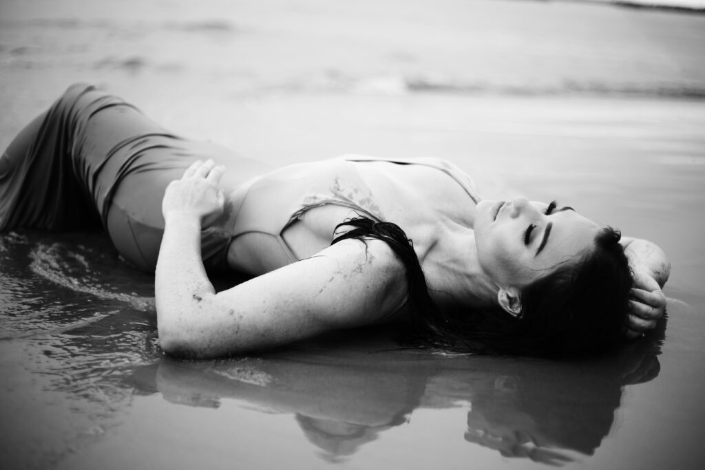 a black and white portrait of a beautiful brunette woman laying in the water at the beach with a wet dress and full face of make up her hands in her hair -bisoux de lune meagan reily