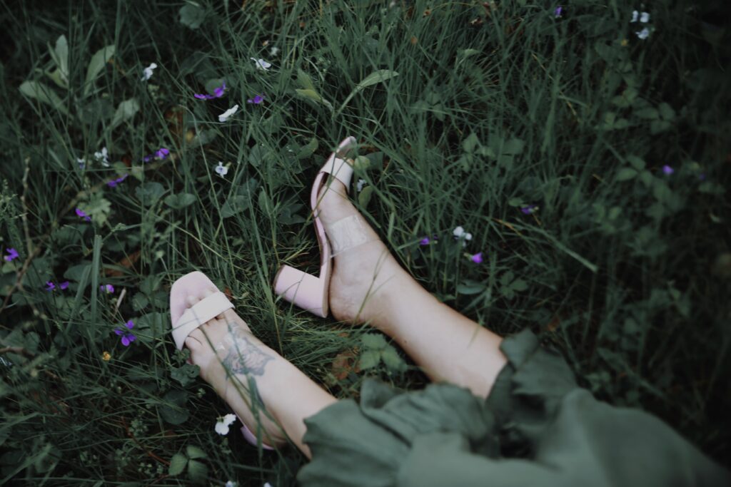 artsy photograph with a lot of greens and hints of purple and white florans of a womans feet in high heels laying in a field in a green dress -bisoux de lune meagan reily