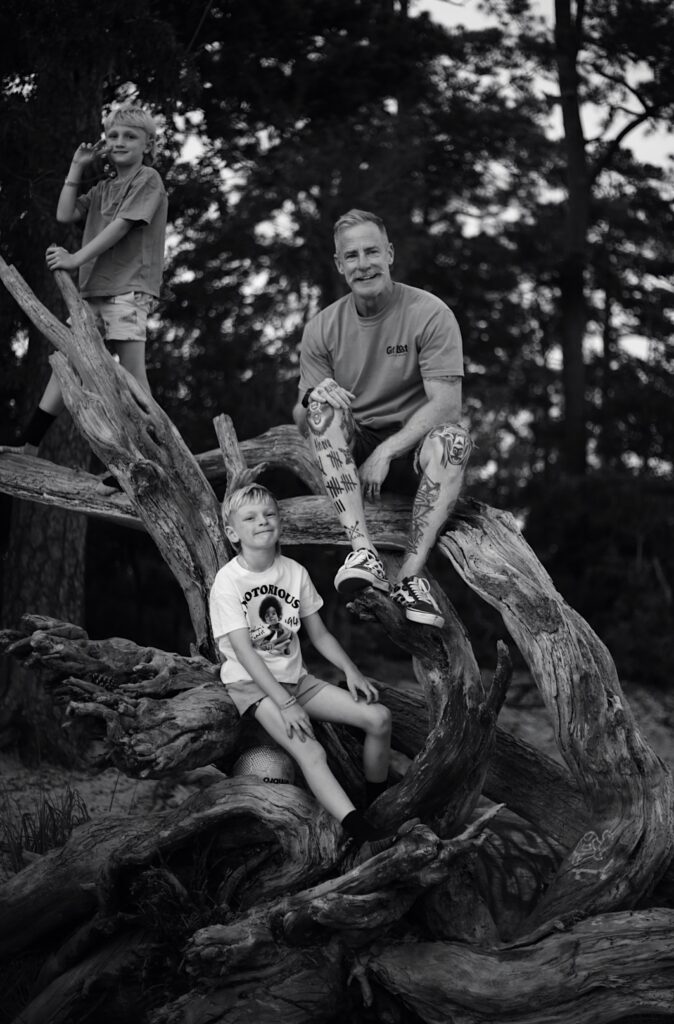 black and white photograph of a heavily tattooed father with a mustache and his two young blonde preteen sons at the state park climbing on a tree and smiling it is a high contrast black and white photograph -bisoux de lune meagan reily