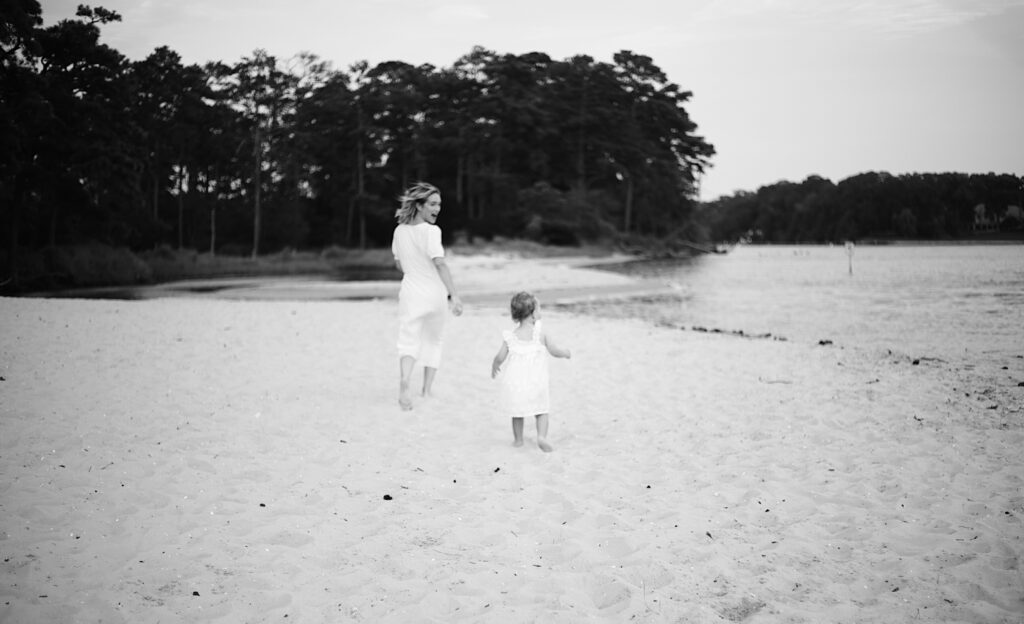 scenic beach shot of young beautiful mother and small baby running on the beach in virginia beach black and white photo with dark trees in the background the image is very whimsical and fun and sentimental there is water in the backgound -bisoux de lune meagan reily