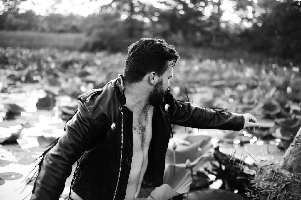 black and white portrait of a bearded musician who is shirtless in a leather jacket jumping out of a lily pond