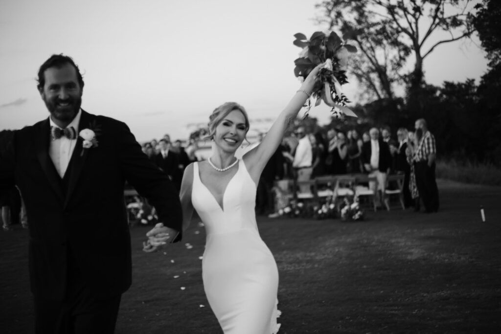 black and white photo of bride and groom after ceremony with bride holding flowers in the air and smiling -bisoux de lune meagan reily