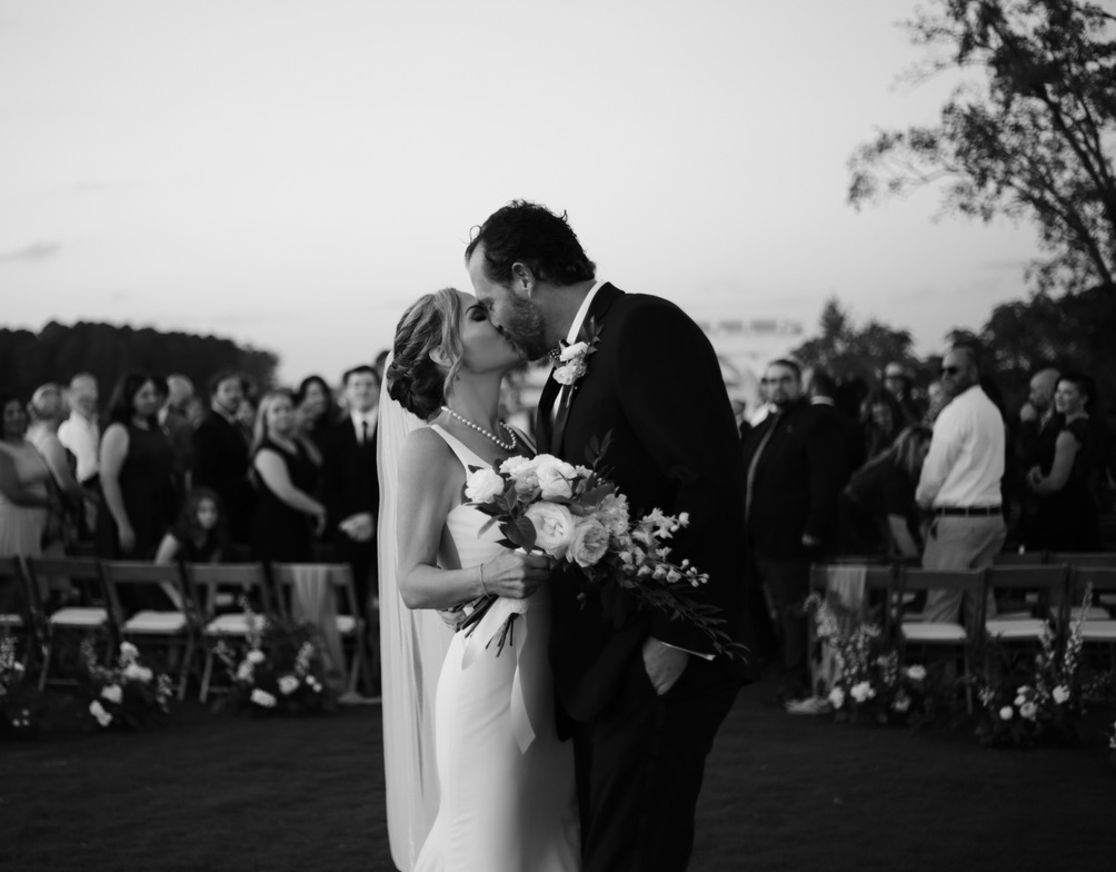 black and white image of bride and groom after their ceremony kissing at the end of the aisle as guests look at them -bisoux de lune meagan reily