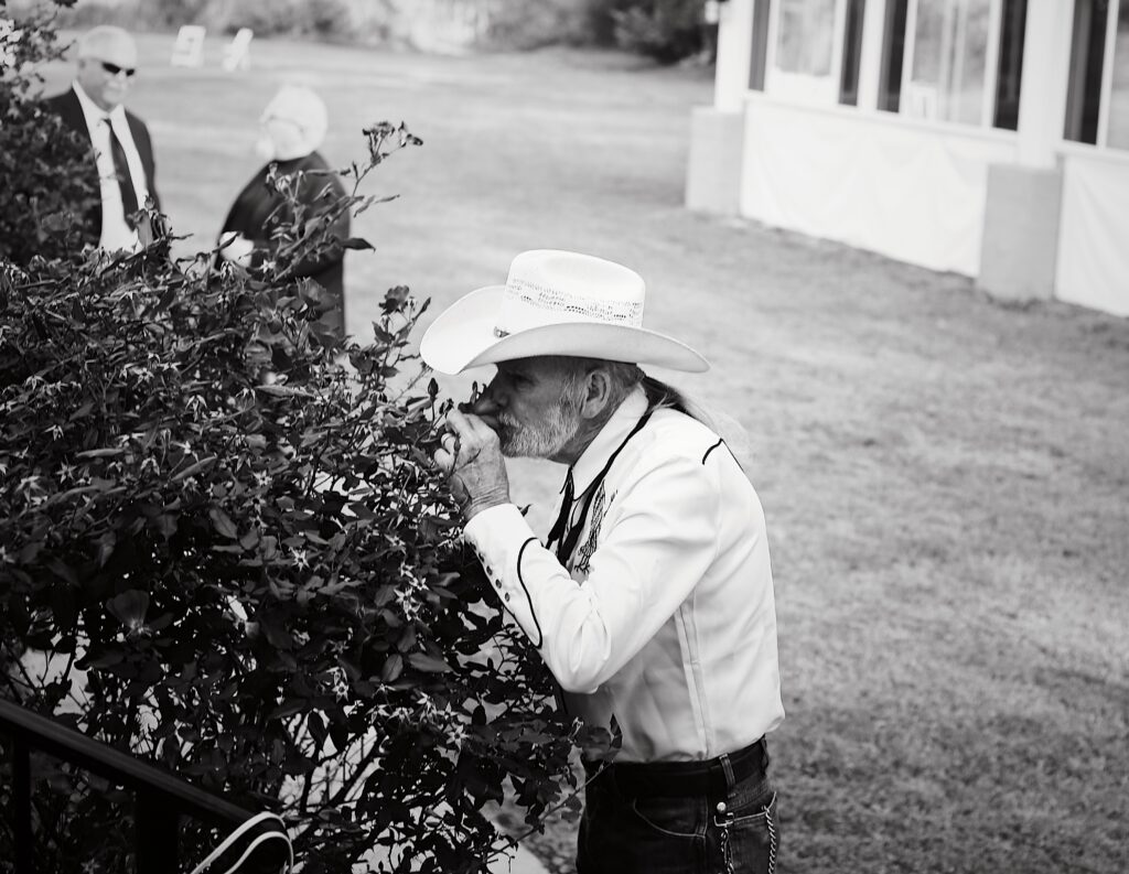 black and white portrait at a wedding of a cowboy dressed up in a white cowboy hat and bolo smelling a rose bush with the flower pressed to his nose -bisoux de lune meagan reily