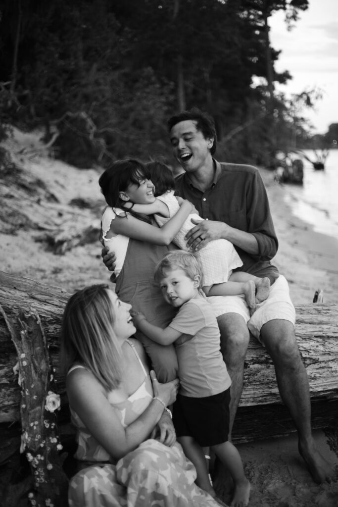 black and white family beach portrait on top of a tree of a mother and father smiling as their three young children hug each other